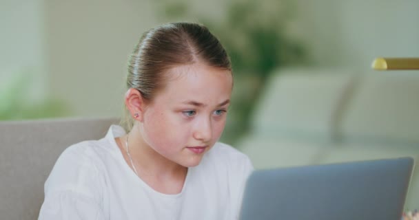 Laptopun önündeki genç kızın yakın plan portresi, ekrana ve kameraya bakıyor ve gülümsüyor. Arka planda bitkiler, kanepe, beyaz duvarlar. — Stok video