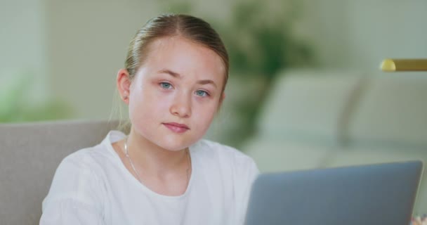 Primer plano retrato adolescente chica delante de la computadora portátil, mira en la pantalla y hacia la cámara, y sonríe Plantas, sofá, paredes blancas en backgroung — Vídeos de Stock