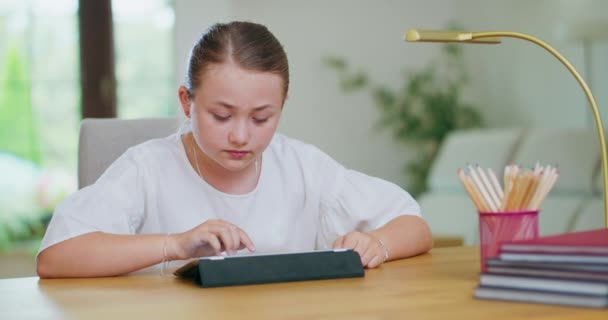 Skupiona nastolatka przy biurku, przewijanie tabletu Książki, ołówki i lampa na pierwszym planie Pierwszy plan i tło są zamazane — Wideo stockowe