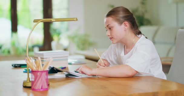 Menina adolescente focada, na mesa, escreve em caderno e pergaminhos tablet Lápis, lâmpada, tablet, cadernos, livros na mesa Paredes, sofá, janela no fundo são borrados Imagem de lado — Vídeo de Stock