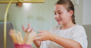 Kapalı odaklı genç kız, masanın başında, dizüstü bilgisayarın önünde, biriyle iletişim kurarak konuşuyor ve el kol hareketleri yapıyor, gülümseyen kalemler, odak dışı lamba bulanıklaşıyor.