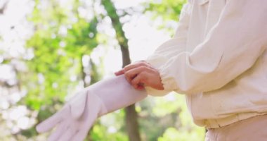 Beyaz koruyucu takım elbiseli erkek arıcının sağ eli beyaz koruyucu eldiven giyer ve eldivenin ellerinin iyi olup olmadığını kontrol eder. Ormandaki ağaçların arasında kalır. Sol el