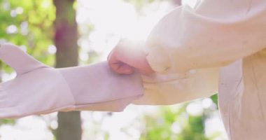 Beyaz koruyucu takım elbiseli erkek arıcının sağ eli beyaz koruyucu eldiven giyer ve eldivenin ellerinin iyi olup olmadığını kontrol eder. Ormandaki ağaçların arasında kalır. Sol el