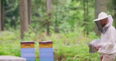 Beyaz koruyucu kıyafetli sakallı genç arı yetiştiricisi, kovanların arasında, ormanın ortasında ağaçlar ve ormandan çıkan dumanla birlikte yeni bir metal içiciyi tutuyor.