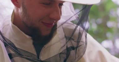 Beyaz koruyucu elbiseli, gülümseyen, ormanın ortasındaki Apiary kovanında bal peteği ve arılarla çerçeveleri inceleyen ve arka planda yeşil ağaçlar olan erkek sakallı arı yetiştiricisinin yakın görüntüsü.
