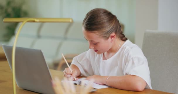 Chica adolescente enfocada cerrada, en el escritorio, hace tareas en el libro de trabajo y las comprueba en el portátil Lápices, lámpara fuera de foco El fondo está borroso — Vídeo de stock