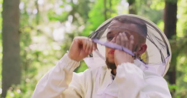 Portrait male bearded handsome beekeeper in white protective suit wears a round protective veil and slightly smiles, staying among the trees in forest Slightly blurred trees in background — Stock Video
