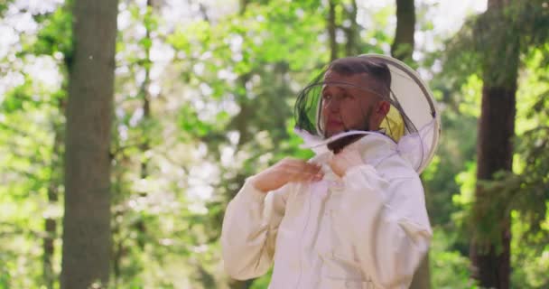 Мужчина бородатый красивый пчеловод пристегивает молнию белого защитного костюма и носит круглую защитную вуаль и слегка улыбается, оставаясь среди деревьев в лесу — стоковое видео