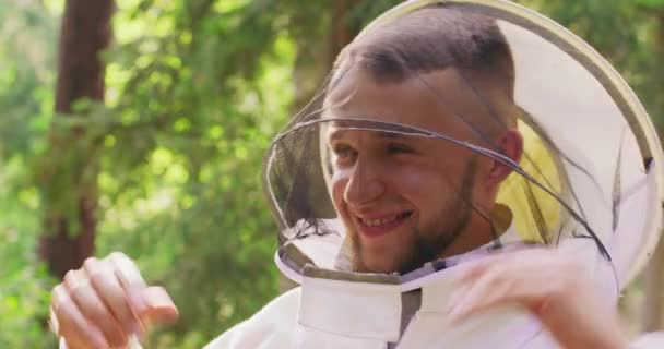 Retrato macho joven barbudo apuesto apicultor en traje protector blanco lleva un velo protector redondo y sonrisas, permaneciendo entre los árboles en el bosque Árboles ligeramente borrosos en el fondo — Vídeo de stock