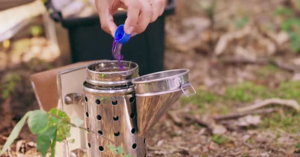 Closeup schijnt nieuwe metalen roker, op de grond in het midden van het bos, en imkers vingers houden fles dop, terwijl hij giet de violette vloeistof fireligther op de houtsnippers in de roker — Stockvideo