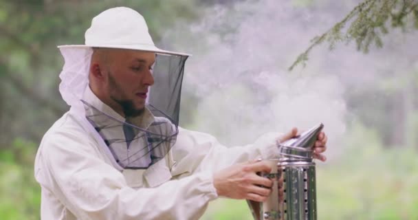 Portrait young, male bearded beekeeper in white protective suit closes a lid of smoking smoker, which is on the hive, and pumps the bellows, what makes smoke go through the lid, then takes the smoker — Stock Video