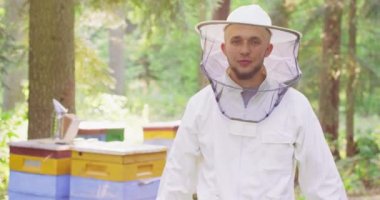 Ormanın ortasında Apiary beyaz koruyucu takım elbiseli ve peçeli genç sakallı yakışıklı arı yetiştiricisi kollarını göğsünde kavuşturup gülümsüyor. İkinci plan:
