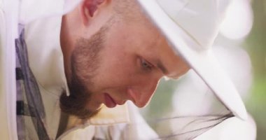 Beyaz koruyucu elbiseli, gülümseyen, ormanın ortasında Apiary 'e bakan genç erkek sakallı arı yetiştiricisinin yakın yüzü bulanıklaşıyor.