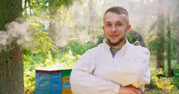 Apiary in the middle of the forest Male young bearded handsome beekeeper in white protective suit crosses his arms on his chest and smiles Second plan: few slightly out of focus hives are among the — Stock Video