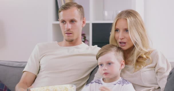 Usmívající se mladá rodina s předškolním synem sedět relaxovat na pohovce sledování filmu v televizi jíst popcorn, šťastní rodiče odpočinku doma s malým dítětem užijte si víkend spolu, bavte se v obývacím pokoji — Stock video