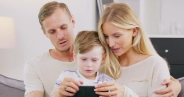 Gülümseyen genç Amerikan ailesi kanepede küçük çocuklarla birlikte oturup tablet üzerinde çizgi film izlerler, mutlu beyaz anne babalar kanepedeki çocukla birlikte rahatlar ve birlikte evde geçirdikleri zamanın videosunun keyfini çıkarırlar.