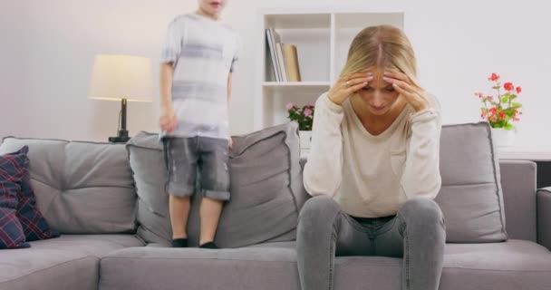Fáradt anya takaró fülek fejfájás, míg a fia ugrál a kanapén otthon szülő hiperaktív gyermek — Stock videók