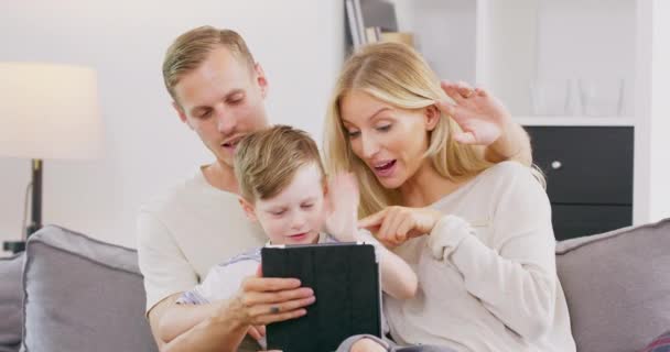 Madre, padre e hijo usando tableta digital haciendo videollamada y saludando Hola sentado en el sofá en la sala de estar — Vídeos de Stock