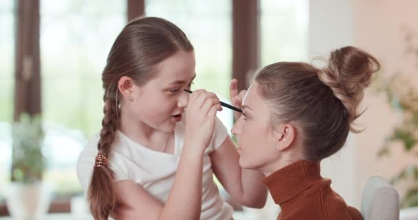Teen girl is learning to apply make up to her mother - she applies the eyshadows on the eyelids of mother, and they are talking, and smiling Window, white wall and green plants at background — Stock Video