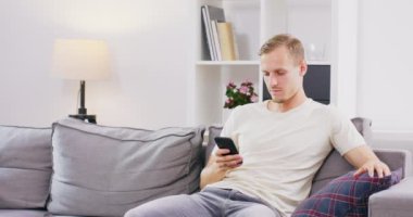 Yakışıklı genç adam akıllı telefondan bilgi topluyor kadın oturma odasındaki kanepede dinlenen kocasını kucaklıyor ve öpüyor ve akıllı telefonuyla interneti karıştırıyor.