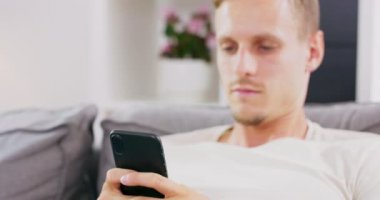Happy Young, Home Man 'de bir koltukta otururken akıllı telefon kullanıyor, interneti kolaçan ediyor, videoları izliyor ve evdeki sosyal ağları kullanıyor