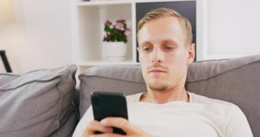 Pozitif düşünceli beyaz adam oturma odasında kanepede oturuyor, akıllı telefon kullanıyor, mesaj yazıyor, haberleri okuyor, internet sitelerinde alışveriş yapıyor, sosyal ağları kaydırıyor.