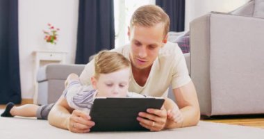 Gülümseyen genç Amerikan ailesi halının üzerine uzanmış çocuk tabletinde çizgi film seyrediyor, mutlu beyaz bir ebeveyn, divanda çocuk ile eğleniyor ve evde birlikte vakit geçirirken videonun keyfini çıkarıyor.