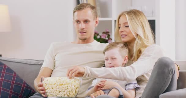 Optimistisk familj i avslappnade kläder sitter på soffan i ljust rum och skrattar medan du tittar på TV och äter välsmakande popcorn under solig dag hemma — Stockvideo