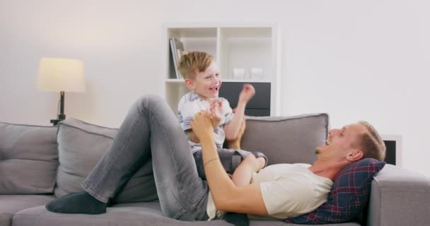 楽しいハンサムなお父さん抱っことくすぐったい笑いの小さな息子ソファでリビングルーム男の子と彼の父親は一緒に遊んでいます家族の楽しい時間の概念 — ストック動画