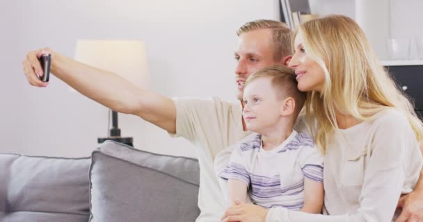 小さな未就学児の男の子を持つ笑顔の若い家族はリビングルームでソファに座って、一緒にセル画を作ります、小さな子供の息子を持つ幸せな親は自宅でスマートフォンで自画自賛 — ストック動画