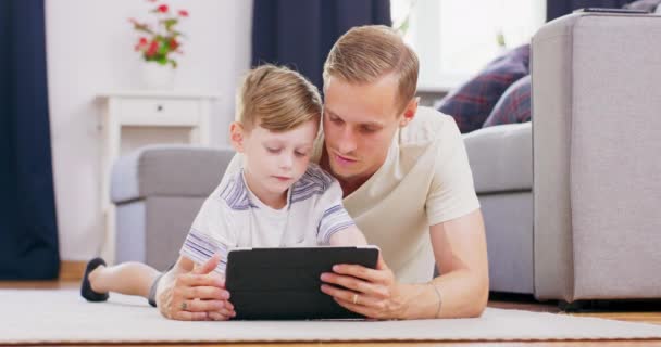 Przystojny mężczyzna i syn w domu leżący na podłodze w salonie i korzystający z tabletu pc Portret wesołego taty i chłopca grających w grę na tablecie cyfrowym relaksujących się na dywanie w domu — Wideo stockowe
