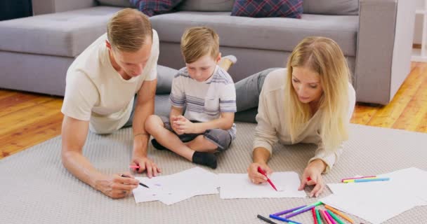 Mignon petit garçon assis sur le sol engagé dans la peinture de l'activité familiale dans l'album avec de jeunes parents, heureux maman aimante et papa se détendre passer du temps avec petit fils dessin image avec des marqueurs de coloration — Video