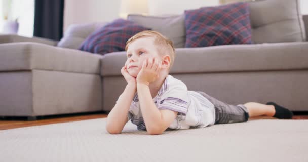 Χαριτωμένο αγοράκι με ξανθά μαλλιά είναι ξαπλωμένο στο πάτωμα κοιτάζοντας το αγαπημένο του καρτούν στην τηλεόραση — Αρχείο Βίντεο
