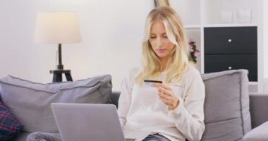 Gülümseyen ve mutlu bir kadın oturma odasındaki koltukta dizüstü bilgisayarla alışveriş yapıyor. Beyaz tenli orta yaşlı bir kız online alışveriş sırasında kredi kartından para harcıyor.