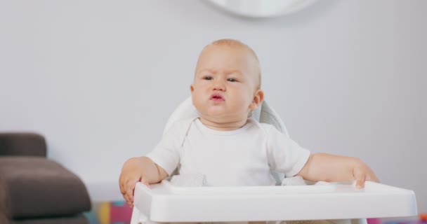Beyaz bebek sandalyesinde komik suratlar yapan ve arka planda beyaz duvarlar çizen bir bebek portresi. — Stok video