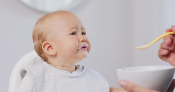 Gros plan du jeune bébé en blanc chaise haute bébé et la main des pères avec une cuillère, alors qu'il nourrit le bébé, qui mange et fait grimaces drôles murs blancs sur fond — Video