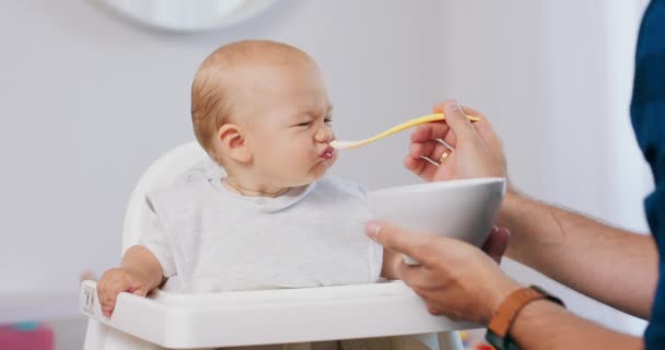 Beyaz bebek sandalyesinde küçük bir bebek ve bir kaşık ve kase ile babasının elleri, bebeğini beslerken ve komik somurtmalar yaparken Baba peçeteyle bebeğin yüzünü siler. — Stok video