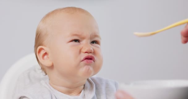 Κοντινό πλάνο του μικρού μωρού σε λευκή παιδική καρέκλα και οι πατέρες χέρι με ένα κουτάλι, ενώ ο ίδιος ταΐζει το μωρό, ο οποίος τρώει και κάνει αστεία γκριμάτσες Λευκοί τοίχοι στο παρασκήνιο — Αρχείο Βίντεο
