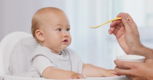 Primer plano del bebé en silla de bebé alta blanca y las manos de los padres con una cuchara y un tazón, mientras que él está alimentando al bebé, que coge la cuchara en la mano de los padres y come haciendo muecas divertidas — Vídeo de stock