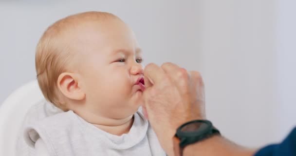 Κοντινό πλάνο του μικρού μωρού σε λευκή καρέκλα και οι πατέρες χέρι με ένα κέικ, ενώ ο ίδιος ταΐζει το μωρό, ο οποίος τρώει κάνοντας αστεία γκριμάτσες Ο πατέρας σκουπίζει το πρόσωπο babys με έναν ιστό — Αρχείο Βίντεο