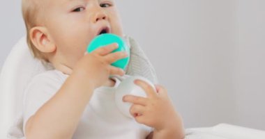 İki elinde yeşil ve beyaz olmak üzere iki plastik top tutan, beyaz bebek koltuğunda oturan ve yeşil olanı ağzına sokmaya çalışan, komik suratını buruşturan bir bebek.