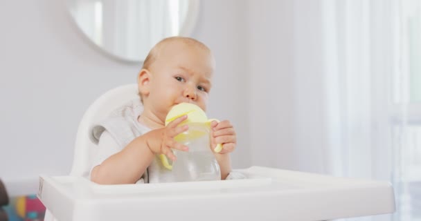 Primer plano del bebé en la silla de bebé alta blanca, que sostiene el biberón de los niños con ambas manos, y trata de beber agua haciendo muecas divertidas — Vídeo de stock