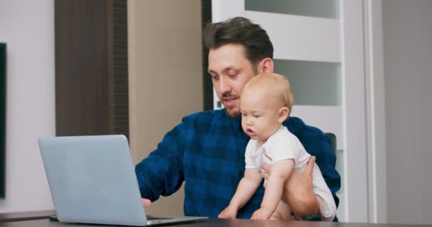 Молодой бородатый мужчина работает за столом дома с ноутбуком, держит милого ребенка на коленях, смотрит на экран Ребенок смотрит на экран, пытается прикоснуться к ноутбуку — стоковое видео