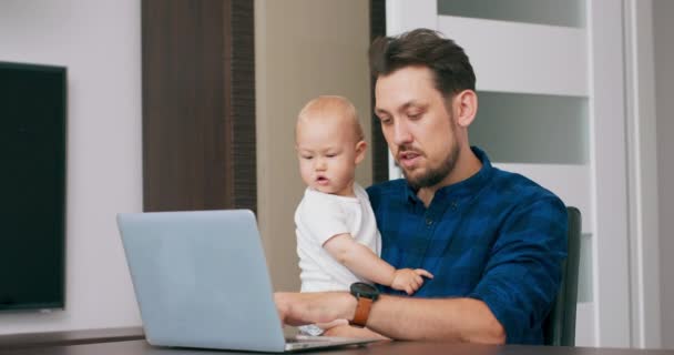 Mladý vousatý muž pracující doma s laptopem, drží roztomilé dítě na kolenou, píše na klávesnici, dívá se na obrazovku Muž mává rukou na pozdrav Dítě se podívá na obrazovku — Stock video