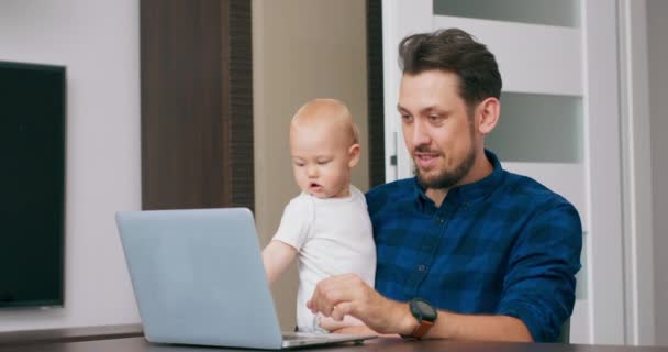 Jonge man met een baard die thuis werkt met een laptop, een schattige baby op de knieën vasthoudt, op het toetsenbord schrijft, naar het scherm kijkt Man zwaait met zijn hand in begroeting Baby kijkt naar het scherm Slow motion — Stockvideo