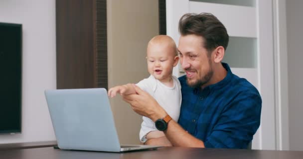 Mladý vousatý muž sedí doma u stolu s laptopem, drží roztomilé dítě na kolenou, mluví přes komunikátor s babys matka Muž mává rukou říká "máma" Dítě se podívá na obrazovku Zpomalený pohyb — Stock video