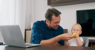 Genç sakallı bir adam dizüstü bilgisayarla evde oturuyor şirin bir bebek bebek bebek sandalyesinde oturuyor, babası bebek ısıran adamı yanağından öpen ipi tutuyor.