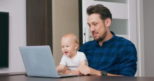 Ung skäggig man sitter vid skrivbordet hemma med laptop, hålla söt baby på knä, prata via kommunikator till babys mor Baby tittar på skärmen, försöker trycka på tangenterna Slow motion — Stockvideo