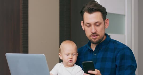 Mladý vousatý muž sedí doma s laptopem, drží roztomilé dítě na kolenou, píše zprávu na smartphone Baby se podívá na smartphone, snaží se ho chytit, pak stiskne klávesnici notebooku — Stock video
