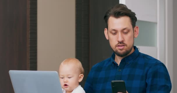 Hombre barbudo joven sentado en el escritorio en casa con el ordenador portátil, sosteniendo bebé lindo en las rodillas, hablando a través del teléfono inteligente El bebé mira el teléfono inteligente, intenta agarrarlo, luego golpea el teclado del ordenador portátil Cámara lenta — Vídeos de Stock